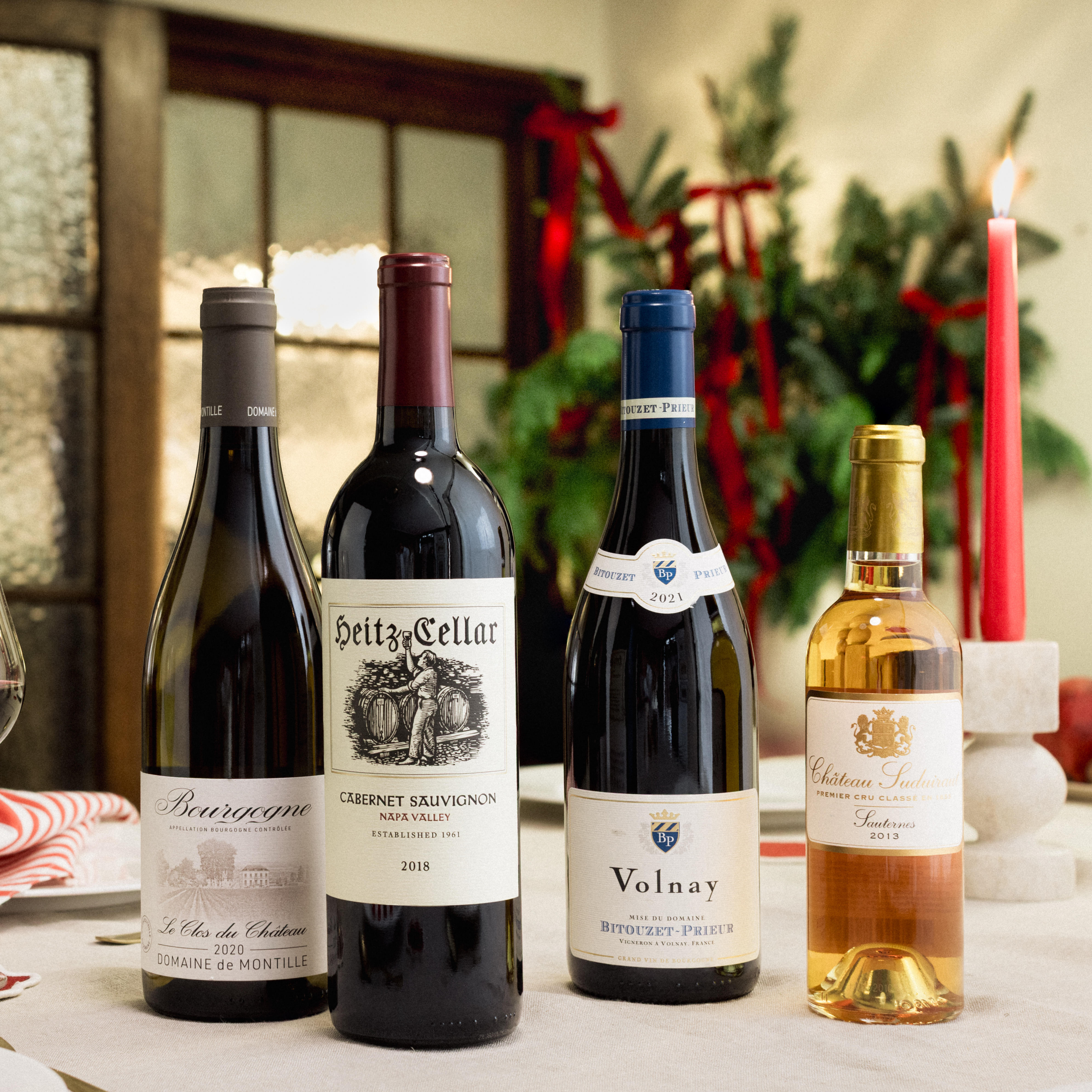 Four bottles of wine are displayed on a festive dining table with seasonal decorations and a red candle. The selection includes Domaine de Montille Baragane, Heitz Cellar Cabernet Sauvignon 2015, Volnay Premier Cru 2012, and Château Suduiraut Sauternes 2013. A glass of red wine and a glass of dessert wine are visible, set against a backdrop of greenery and red ribbons, creating an elegant Christmas setting.